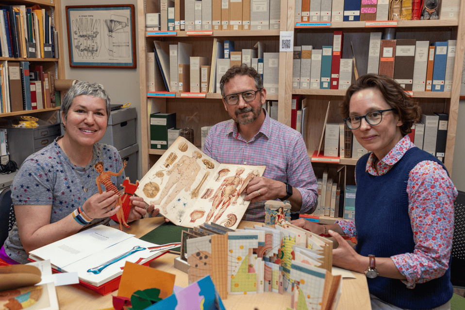 Three people sit at a table which is covered in different types of pop-up books and paper dolls.