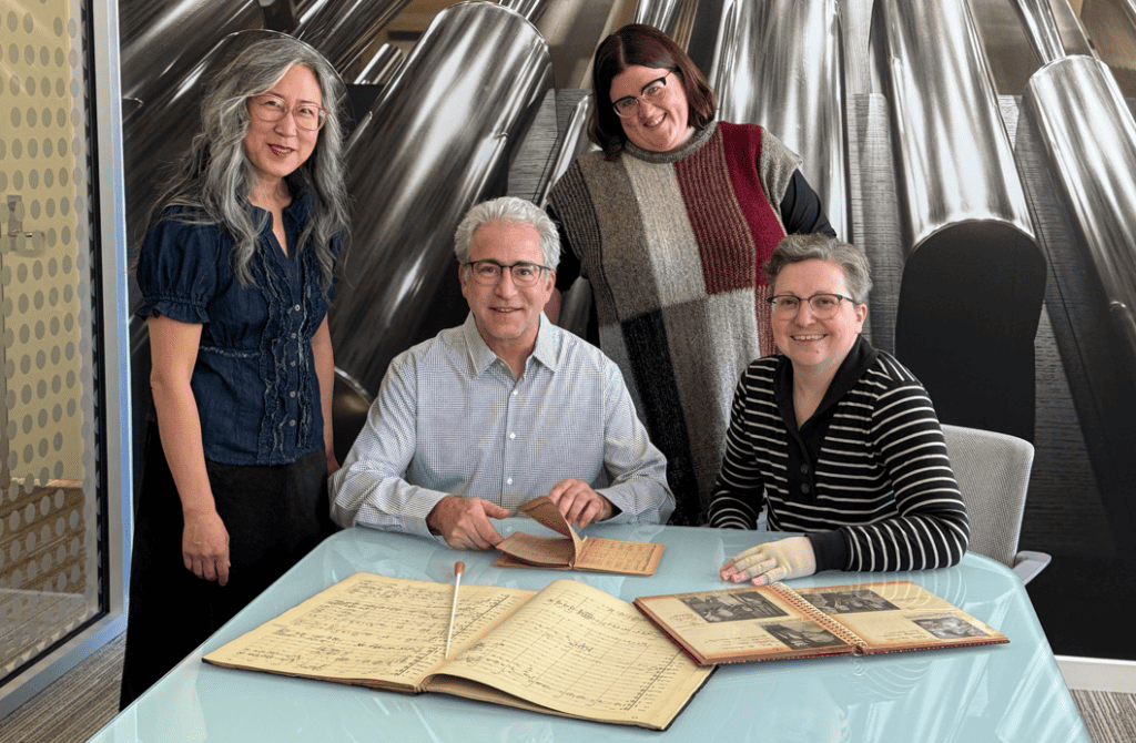 Miera Kim, Carey Bostian, Sarah Suhadolnik, and Katie Buehner pose together behind a table displaying scrapbooks and a conducting baton.