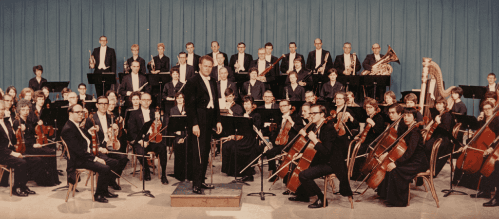 A symphony orchestra is seated together on stage. All are wearing formal attire. The conductor stands on his podium and is turned toward the audience.