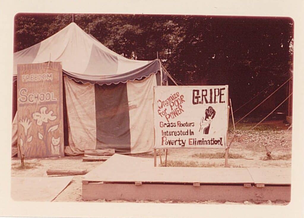 color slide of a striped tent with a hand-painted sign in front of it reading Freedom School; another sign features a white open hand alongside a black fist and reads (G-R-I-P-E: Grass rooters interested in poverty elimination 