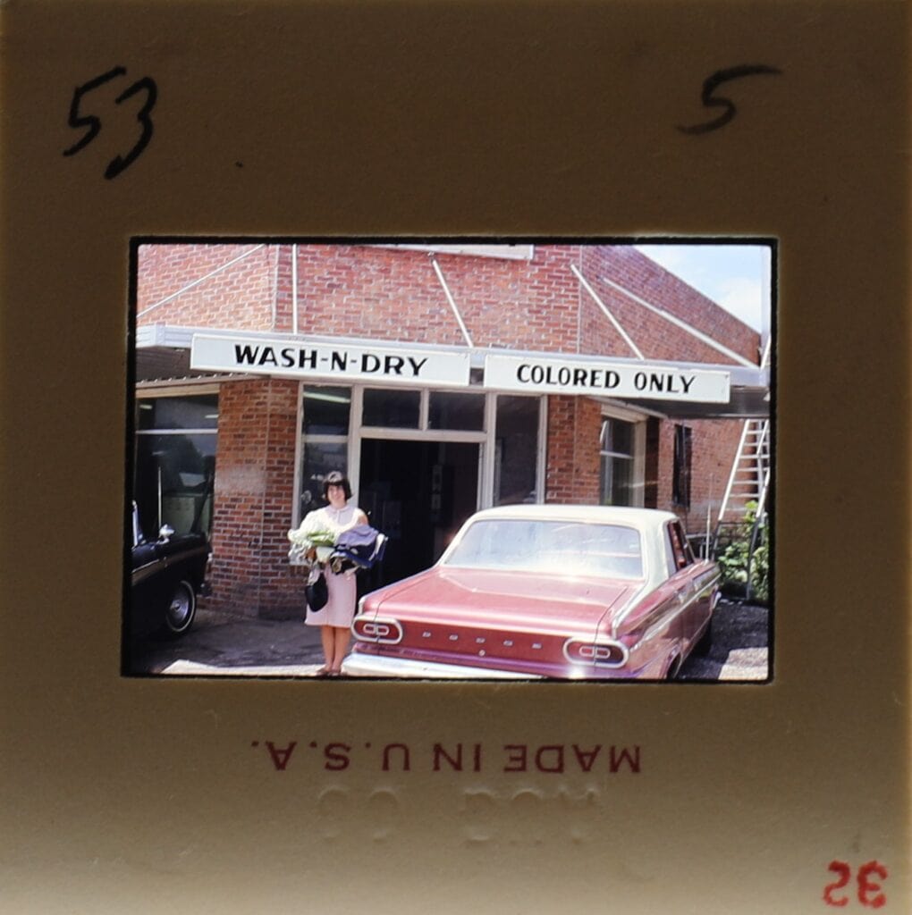 Color slide showing a white woman with arms full of clothes in front of a building with sign reading Wash-N-Dry, Colored Only