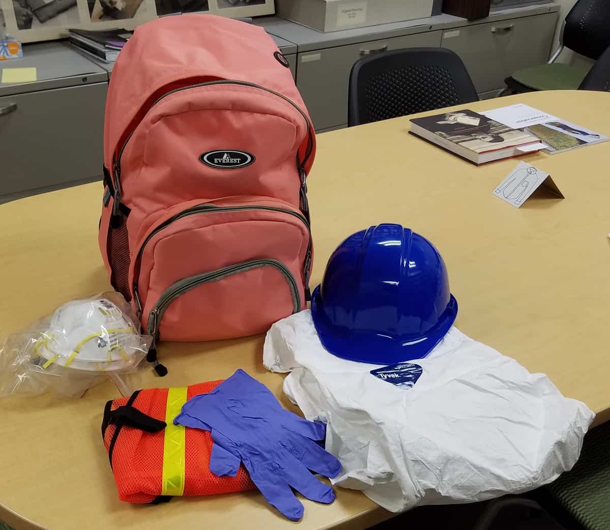 Pictures shows a pink backpack with items sitting in front of it. These items are a hard hat, Tyvek suits, gloves, a safety vest, and respiratory masks.