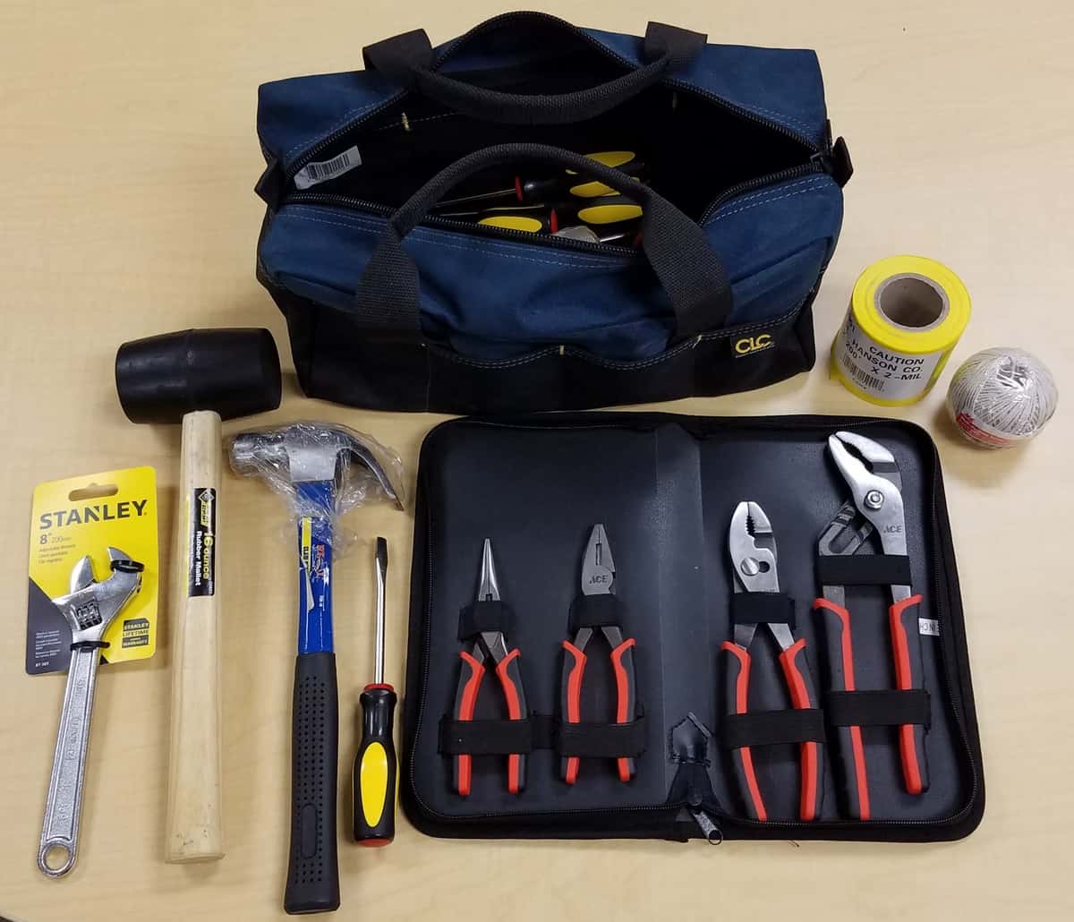 Pictured is a blue bag containing tools with items set out in front of it including a wrench, mallet, hammer, screwdriver, pliers, caution tape, and wire (left to right).