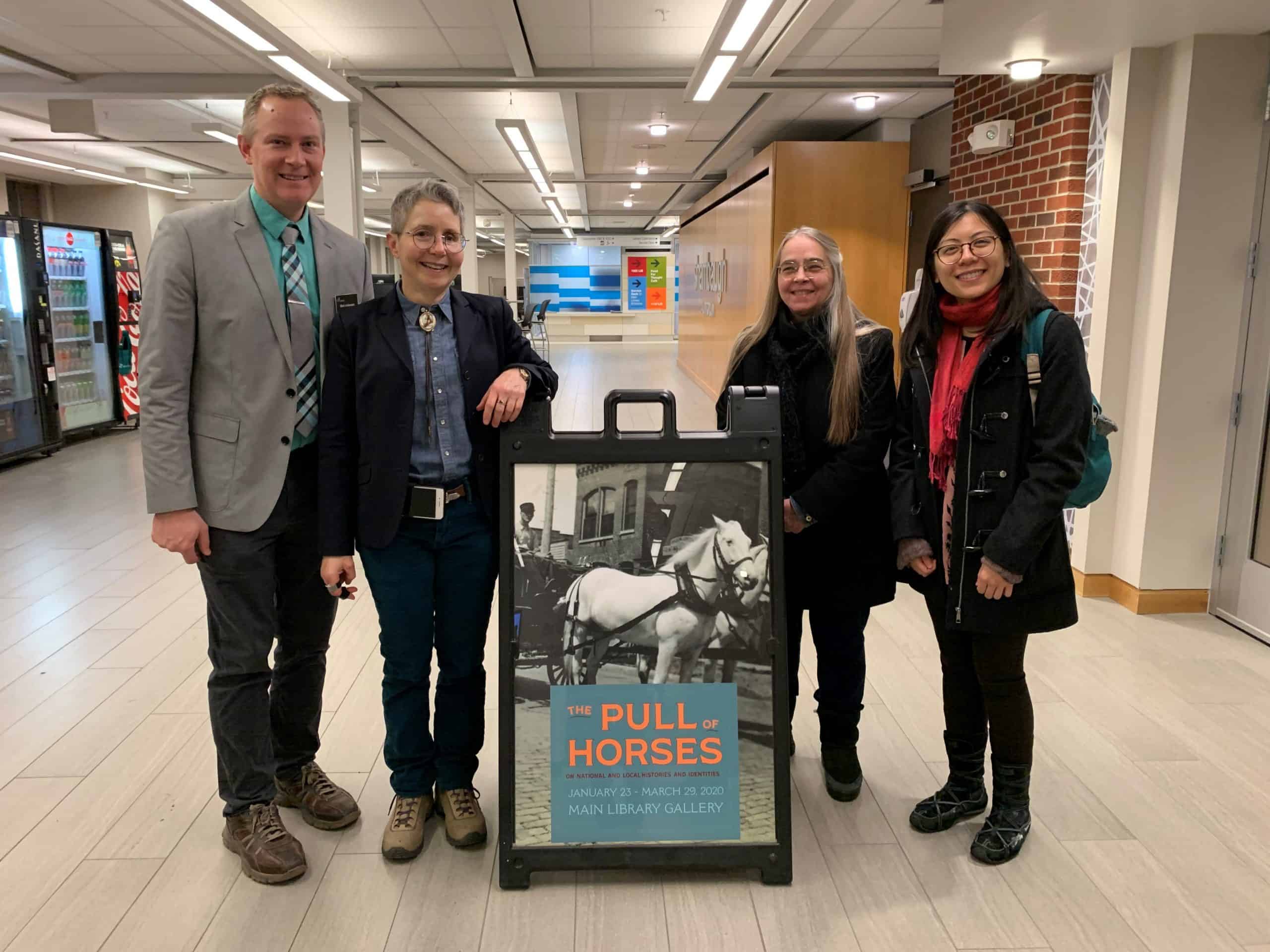 Mark Anderson, Kim Marra, Mary Bennett, and Hang Nguyen stand in a row, smilling, next to a poster of two white horses.
