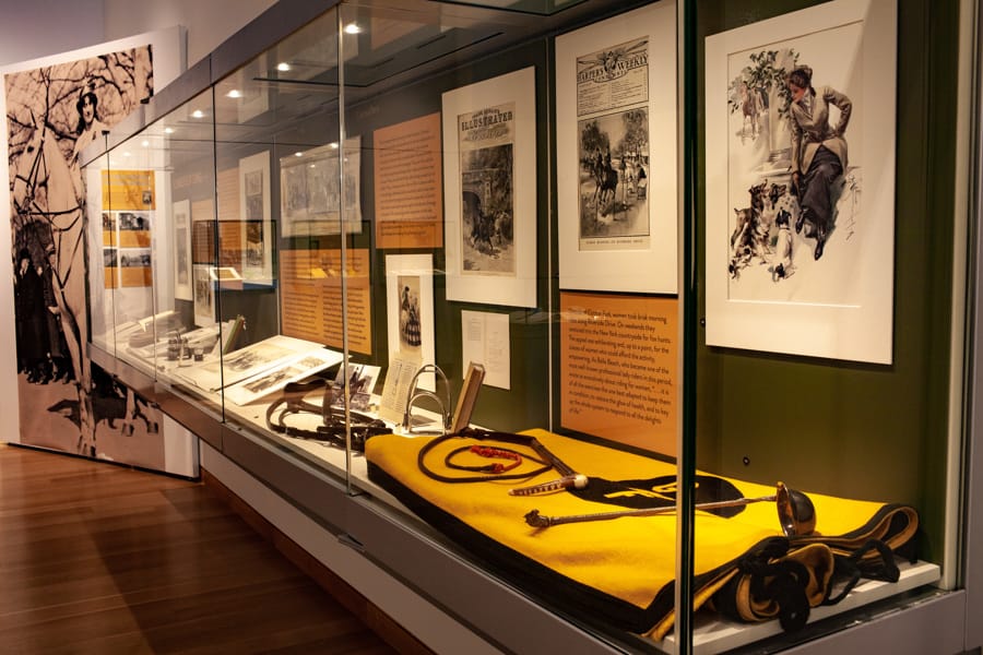 A glass case shows illustrations and photos of women on horseback, and a yellow horse blanket and tack.