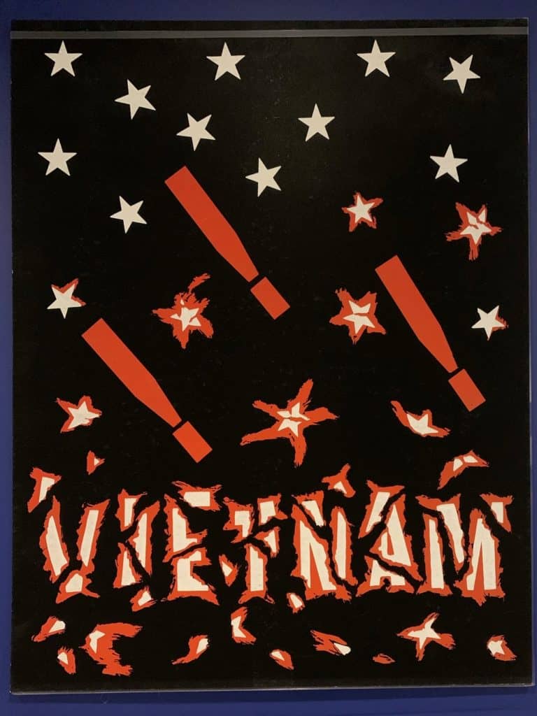 Poster with black background punctuated by falling white stars, small red explosions, and three bombs that look like exclamation points. At the bottom the text says Vietnam and appears wrapped in barbed wire.