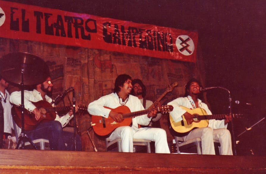 Five Latinx men in white shirts smile, sing, and play guitar on stage. A banner behind them says El Teatro Campesino.