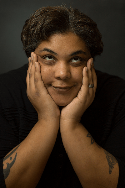 Black author Roxane Gay cups her face between her hands, elbows on the table in front of her. She looks up and she smiles, lips together.