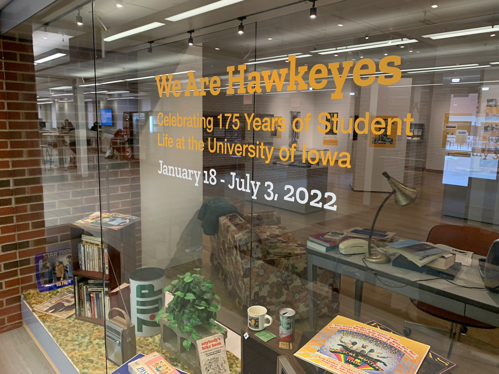 A window display is filled with vintage books, games, records, a record player, green and orange shag carpet, and more to evoke a 1970s dorm room feel. The title of the exhibit is in vinyl on the glass window.