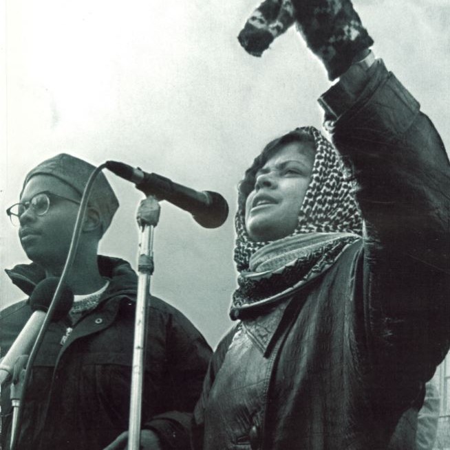 Two students stand at a microphone outside. The photo is in black and white and they are protesting. No context is available as to what they are protesting.