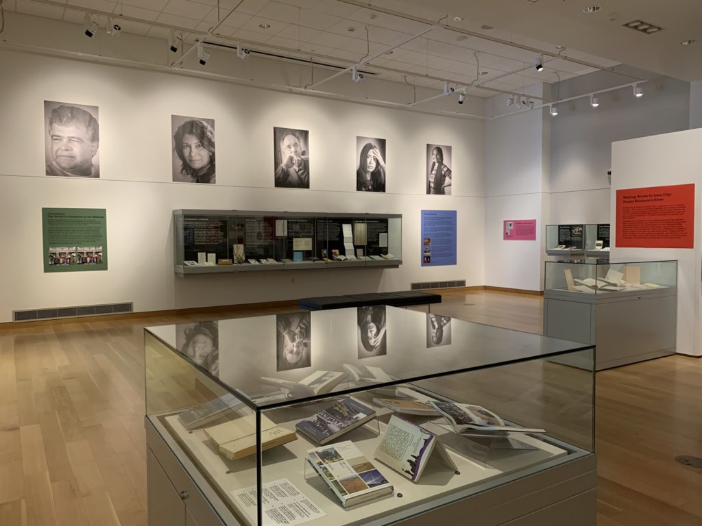 A view of half the gallery includes three display cases filled with books and items at a distance. Colorful text panels are on the walls but the words are indistinguishable. Five black and white portraits of writers from around the world are high up on a white wall above a case.
