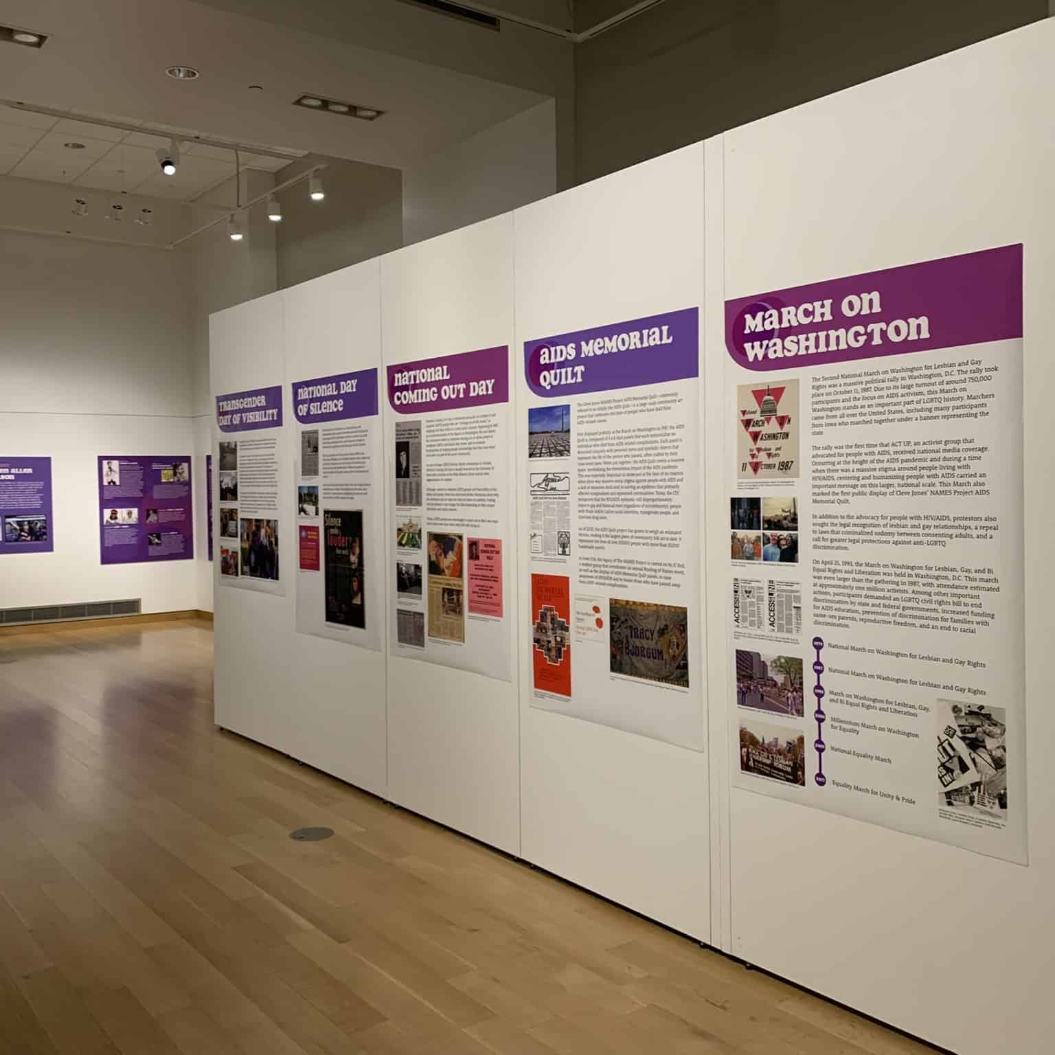 A series of posters are on a large white wall. The posters have headlines like March on Washington, National Coming Out Day, Transgender Day of Visibility, National Day of Silence.