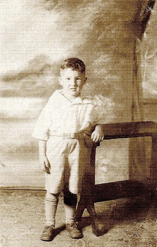 A sepia photo of a young white boy with short hair. He is wearing a simple shirt, trousers, stockings, and shoes. He leans against a table in a photo studio and looks slightly concerned.