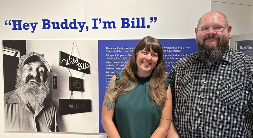 Jen and Brad stand in front of the introductory panel for the exhibit Hey Buddy, I'm Bill. The title is printed on the wall above a black and white photo of Bill, an older white man with a beard who looks joyfully at the camera. He is wearing an engineer's cap. Jen is a white women with long brown hair and is wearing a green top. Brad is a bald white man with a beard and glasses and is wearing a plaid shirt. Both are smiling. It is their first time visiting the completed exhibit.