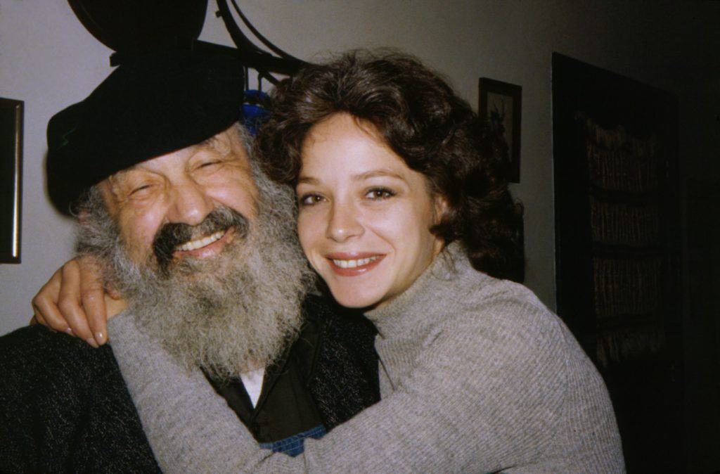 Bev Morrow and Bill Sackter pose together for a photo. Bev, a young women in her 20s with short dark hair, hugs Bill. Both smile. Bill has a gray beard and wears a black cap.