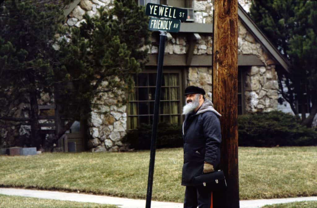 Bill is an older white man with a long gray beard. He wears a black newsboy cap, a dark blue coat, brown gloves, and holds a black metal lunchbox. He is standing next to the intersection of Yewell St and Friendly Ave, indicated by street signs. Behind him is a Moffett House with a stone facade. 