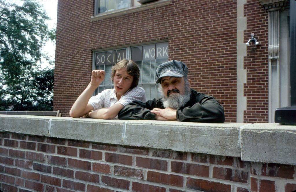 Bill stands with his friend Barry, a 30-something white man. They lean against a brick wall in front of North Hall, another brick building that shows the words Social Work in a window.