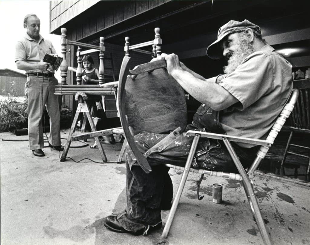 Tom Walz is in the far background of this photo, and in the foreground is Bill sitting in a lawn chair as he works on sanding a wooden chair. Tom and Bill are refinishing furniture together.