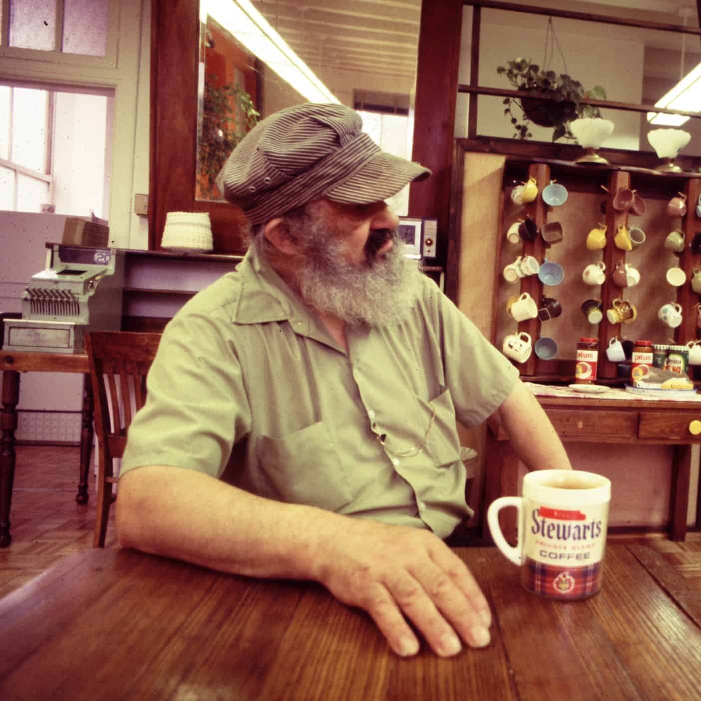 Bill sits in his coffee shop at a table with a cup of coffee. He wears an engineer's cap and suspenders.