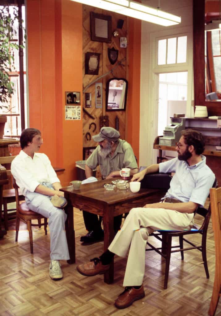 Barry, Bill, and their friend Rabbi Jeff sit together around a small wooden table in Wild Bill's. It is the 1970s. They each have a cup of coffee. The coffee shop has orange walls, many plants, parquet flooring, and lots of antique tools, mirrors, and advertisements on the walls. Several of Bill's award plaques are also displayed in the background