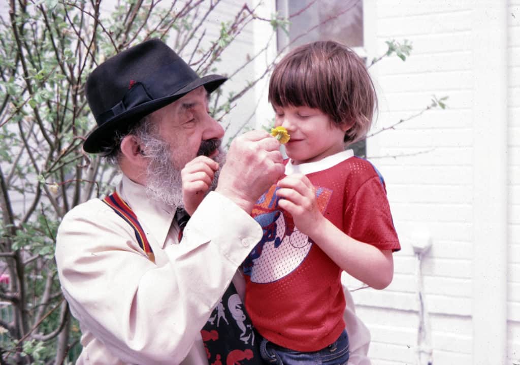 Bill is an older white man with a gray beard. He wears a black fedora, colorful suspenders, and a button up shirt with tie. He is next to a young boy named Clay Morrow and holds a flower out for Clay to smell. Clay smiles with closed eyes as he smells the flower.