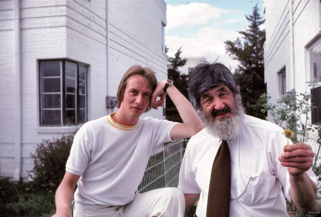 Barry and Bill pose together for a photo. Barry is a young white man in his late 20s. He smiles and rests his elbow on Bill's shoulder. Bill is an older man with a gray beard and he smiles at the camera while holding out a small flower.