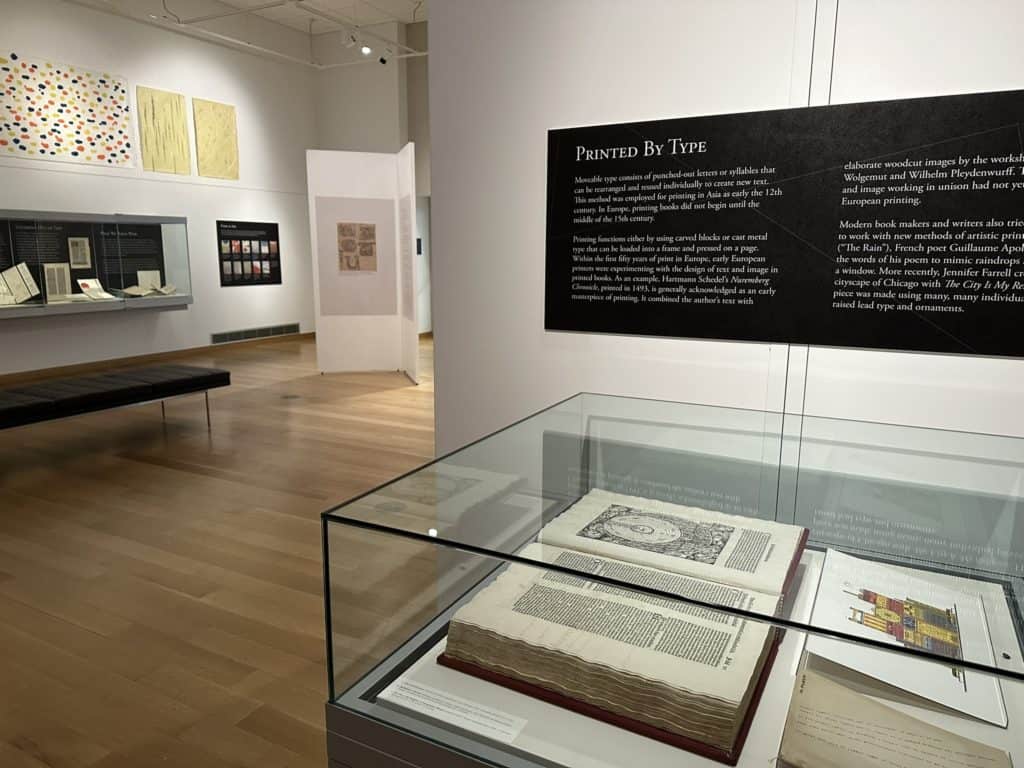 An expansive shot of the gallery, including a large copy of the Nuremburg Chronicle in the foreground in a display case. Beyond, there are many book objects in display cases and large colorful handmade papers on the walls.