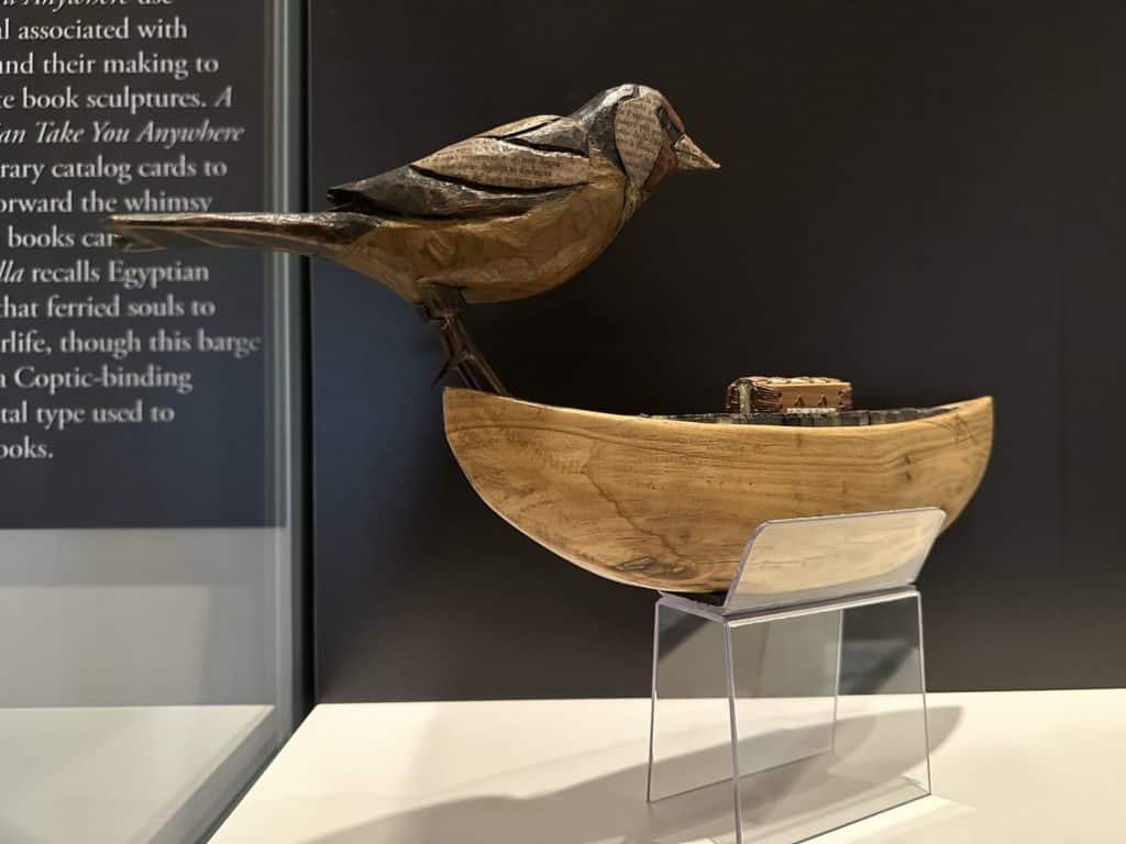 A sculpture of a large bird perched on a bow of a small Egyptian boat. Inside the boat are pieces of metal print type and a very small book with a coptic binding.