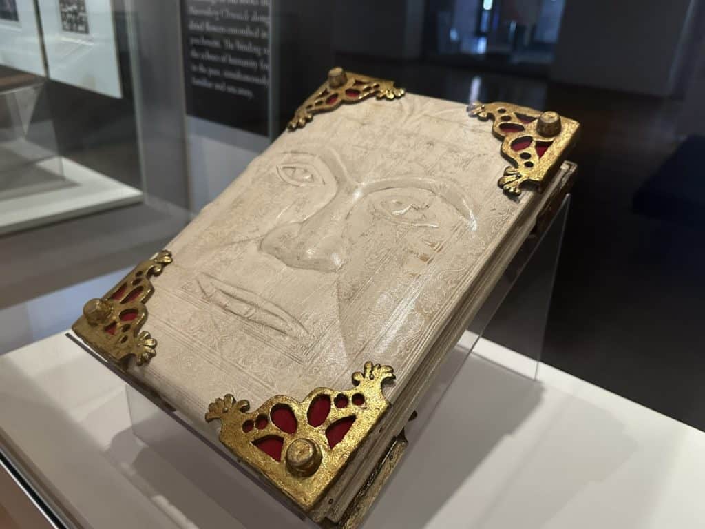 A white leather book with a haunting face in relief on the front cover. Each corner is capped in ornate gold and red. 