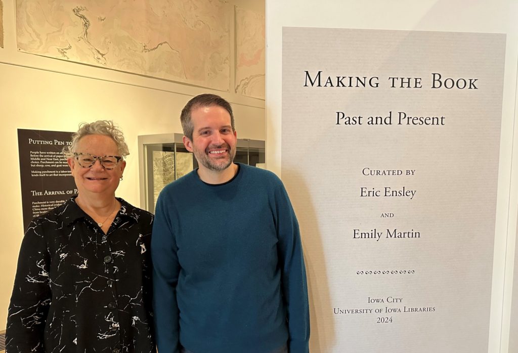 Two people, the exhibit curators, stand in front of a sign that says Making the Book Past and Present, curated by Eric Ensley and Emily Martin, 2024. Emily is a 70-something artist with short gray hair and wears black glasses and a black shirt. Eric is a 30-something librarian with short brown hair and wears a teal sweater. They are both smiling. 