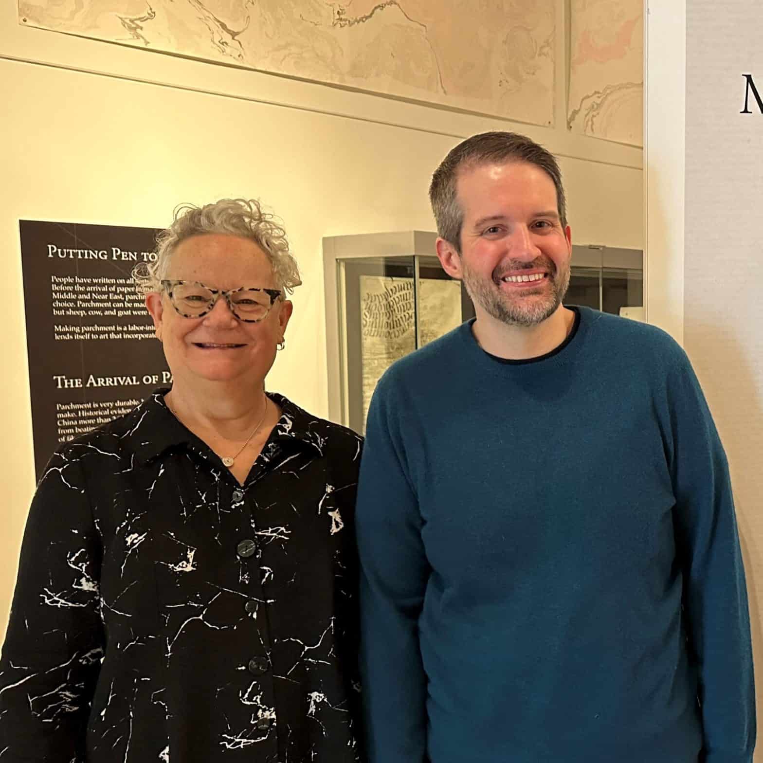 Two people, the exhibit curators, stand in front of a sign that says Making the Book Past and Present, curated by Eric Ensley and Emily Martin, 2024. Emily is a 70-something artist with short gray hair and wears black glasses and a black shirt. Eric is a 30-something librarian with short brown hair and wears a teal sweater. They are both smiling.