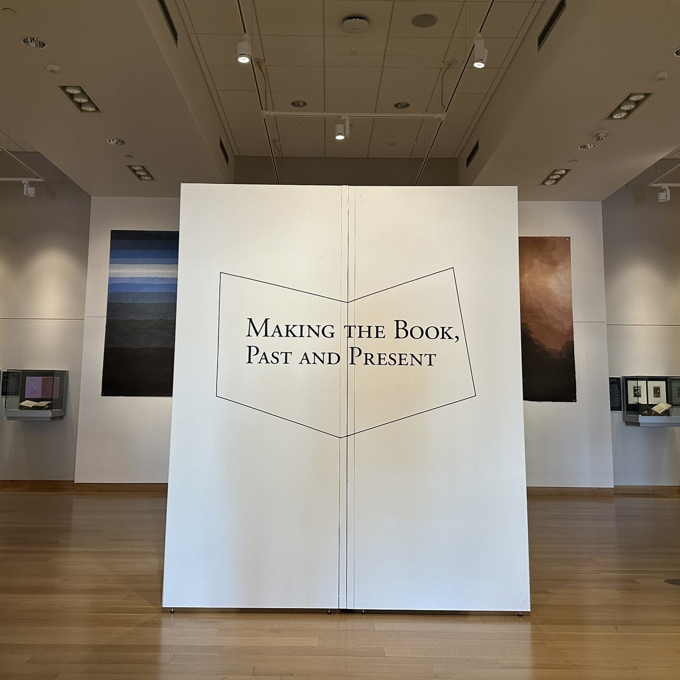 A spacious gallery with two white portable walls in the center, connected. The face of the walls gives the exhibit title of Making the Book, Past and Present in black vinyl.
