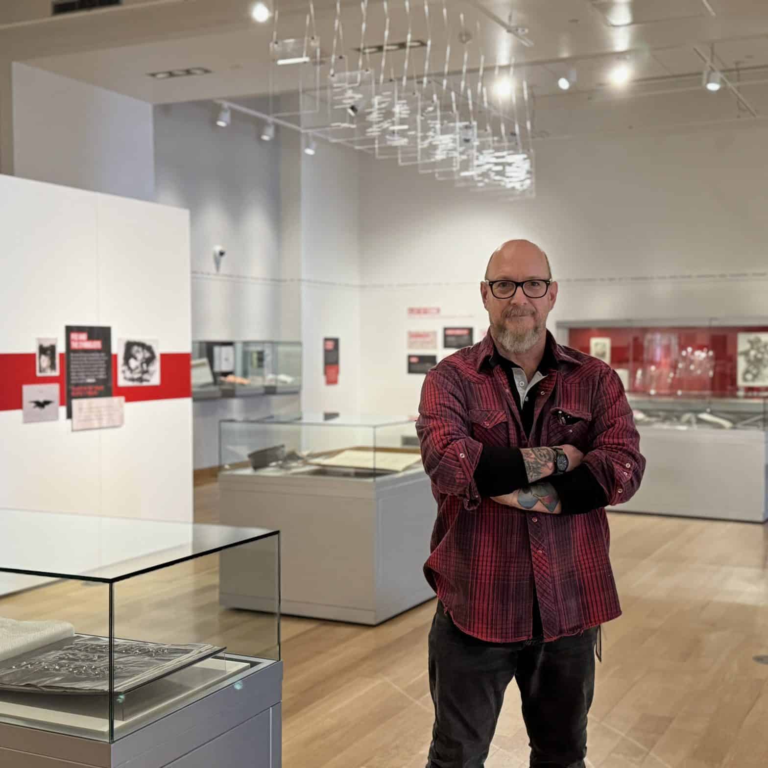 Curator Rich Dana stands in the middle of an art gallery filled with book arts and a plexiglass art piece hanging from the ceiling.