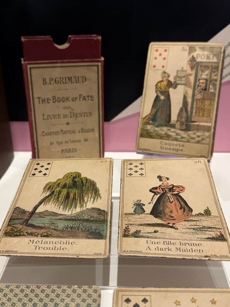 Examples of cards in the card game Livre du Destin. Two illustrated cards are in the foreground. One features a weeping willow and says "trouble." The other features a young woman with brown hair wearing a pink Georgian-style dress. The caption for this card says "a dark maiden."
