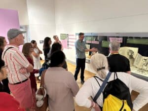 Curator Damien Ihrig stands in front of a display case and points at objects within. He is giving a guided tour to a group, and about 20 people are standing in the exhibit gallery listening to him speak.
