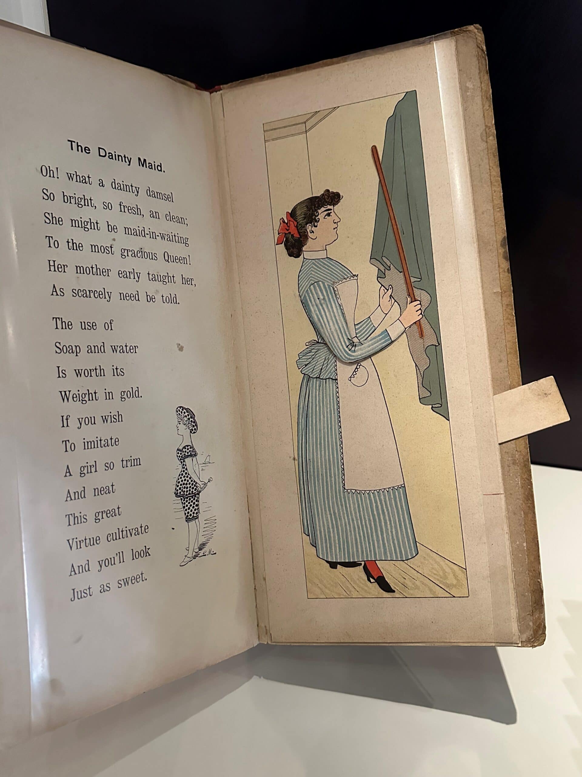 An illustration of a maid wearing a blue and white striped dress and white apron. She is beating dust out of curtains with a rod. The poem next to her says: The Dainty Maid. Oh what a dainty damsel. So bright, so fresh, an clean. She might be the maid-in-waiting to the most gracious Queen! Her mother early taught her, as scarcely need be told. The use of soap and water is worth its weight in gold. If you wish to imitate a girl so trim and neat, this great virtual cultivate and you'll look just as sweet.