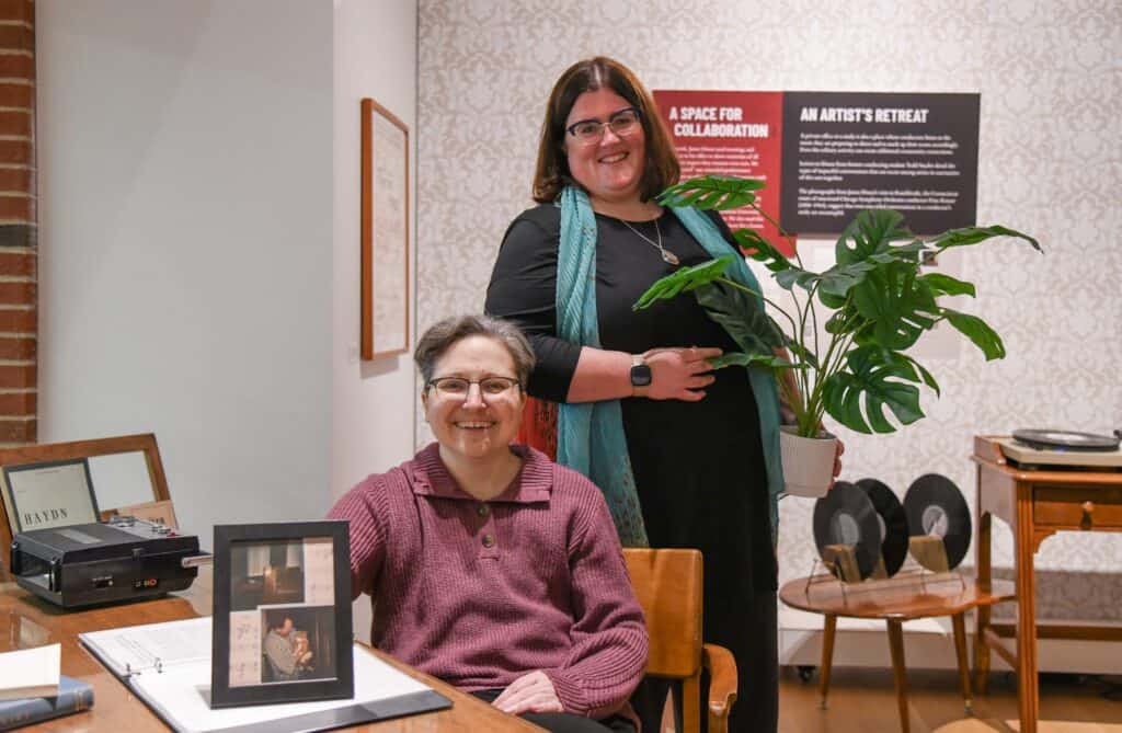 Curator Katie sits in a chair at a vintage desk and curator Sarah stands behind her. They are both smiling, Katie is holding a photo of James Dixon and his cat. Sarah is holding a faux plant. They are at the opening event for their exhibit.