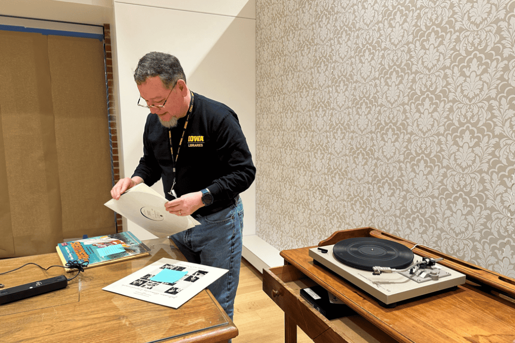 Jose Jimenez takes a record out of a sleeve. He is standing next to a turntable, a disconnected speaker, and a desk as he works to assemble a special audio interactive.