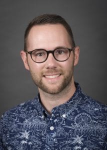 picture of white man with glasses, beard and mustache in blue and white shirt on gray background