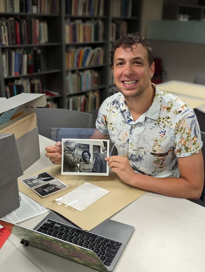 A man sitting at a table with archival boxes and a laptop, holding up a black and white image of a woman