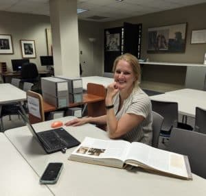Blonde white woman smiling, seated at a desk with a book and laptop next to archival boxes