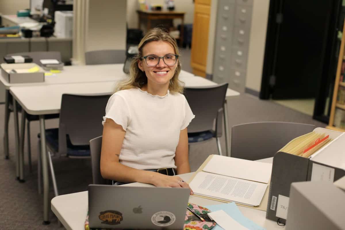 Woman in white shirt sits at table with a laptop and archival boxes