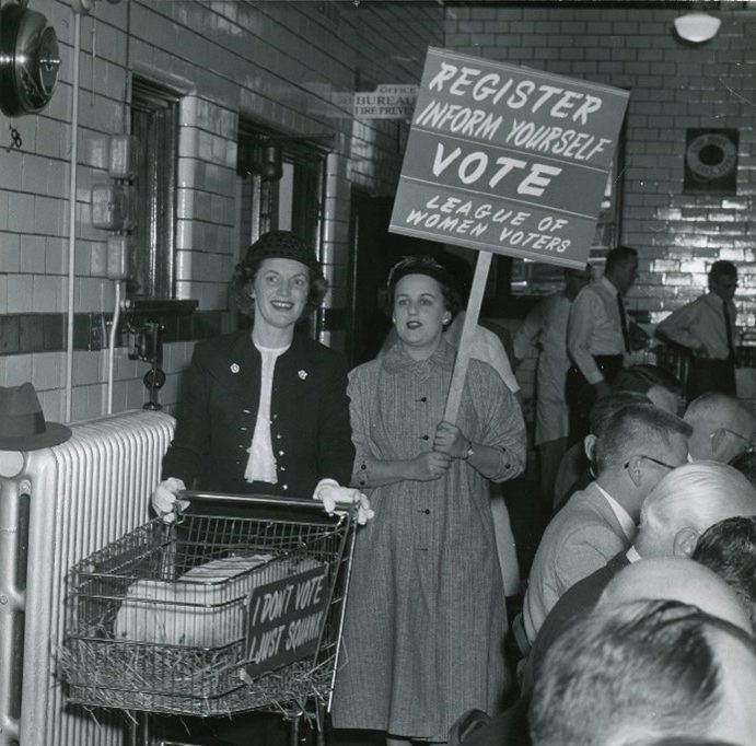 Two women with a shopping cart holding a chicken, carrying a placard encouraging voter registration