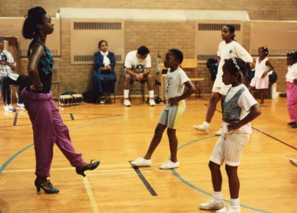 Black woman in athletic clothing and dance shoes demonstrating a dance move to a group of children who are imitating her