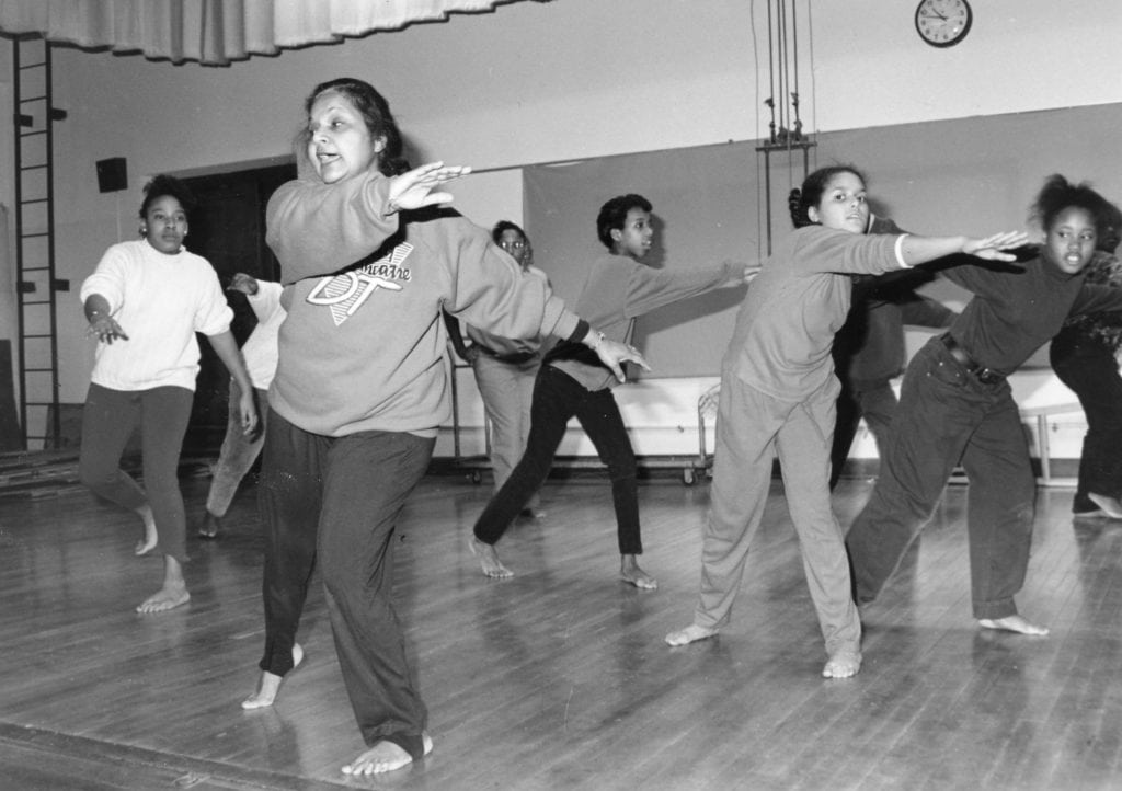 Black and white photograph of a dance class in a gymnasium. The teacher is front and center with several students dancing behind her.