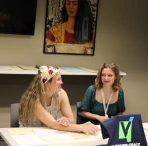 two young women sitting at a table with papers in front of them, smiling at one another