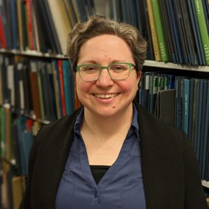 photo of Katie Buehner, white woman, green glasses, in front of library shelves