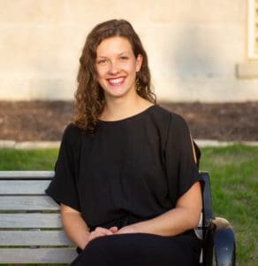 Charlotte is smiling at the camera, sitting on a park bench in a black dress. 