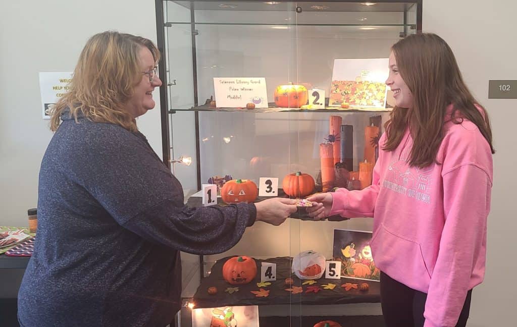one woman handing another woman a giftcard in front of a pumpkin display