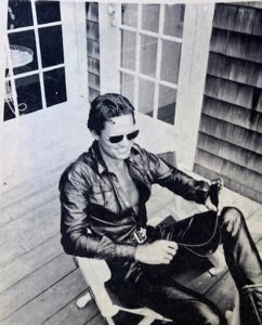 Black and white photo of a young man sitting in a chair, wearing all leather and sunglasses